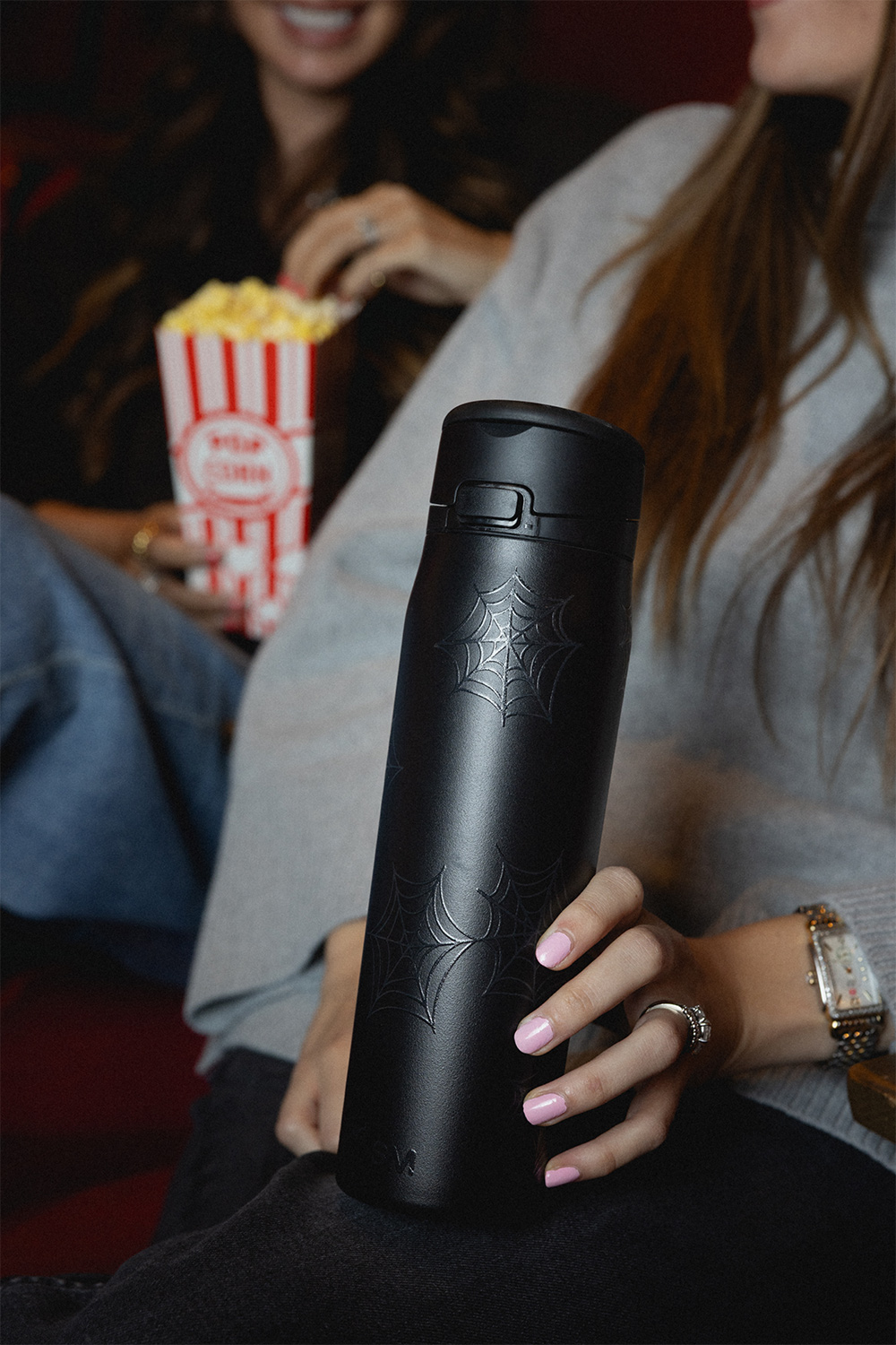 Person holding a black bottle with spider web design, another holding popcorn.