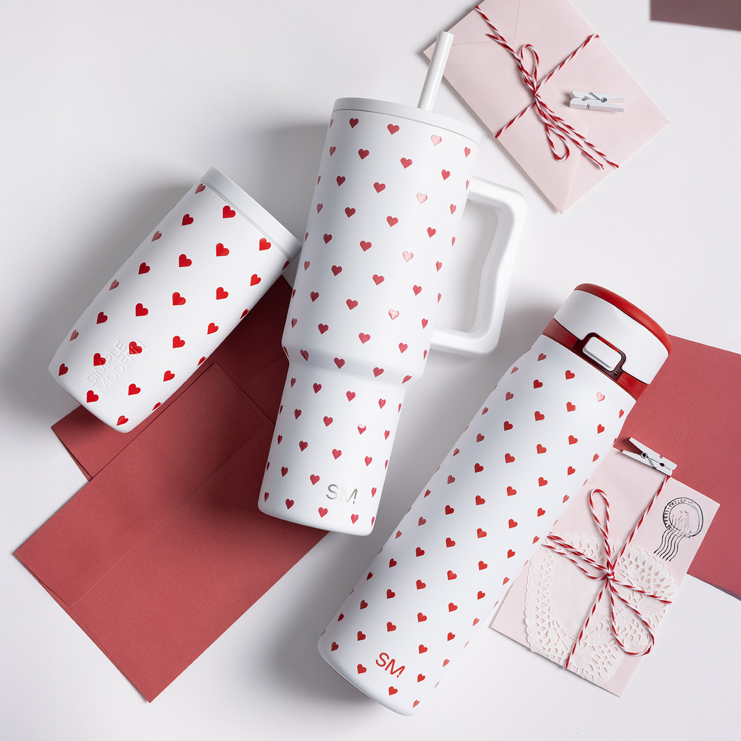 An overhead view of three white travel mugs with red heart patterns, arranged with red and pink envelopes.