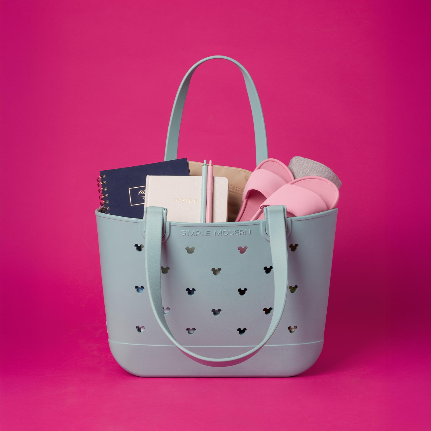 Blue tote bag with notebooks, pink sandals, and socks on a pink background.