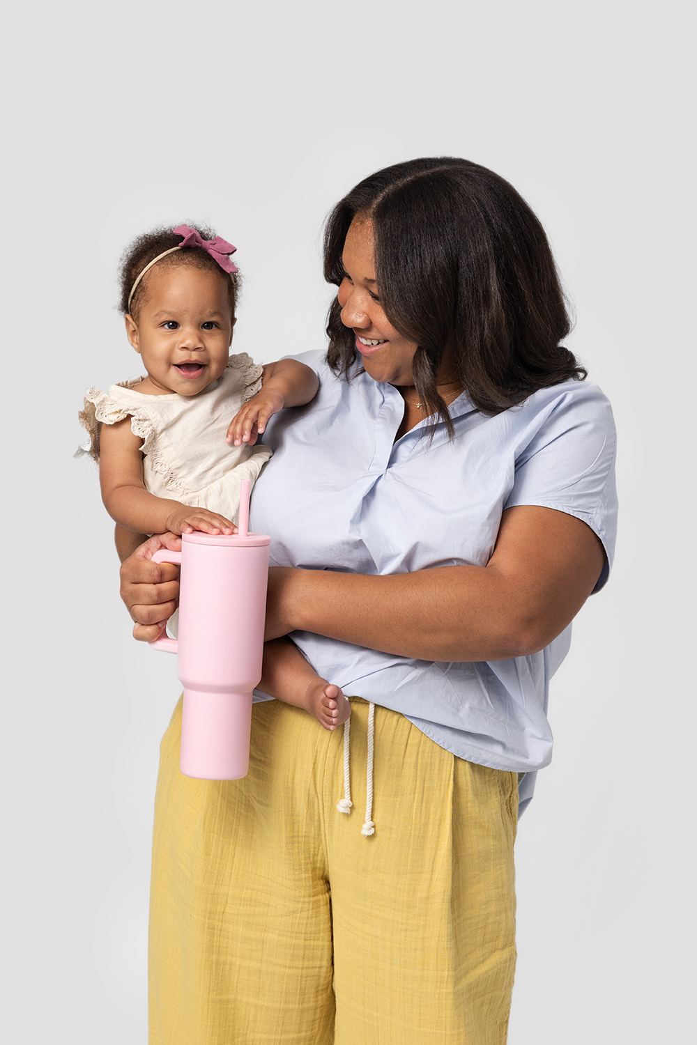 A smiling woman holds a baby and a large pink tumbler against a plain background.