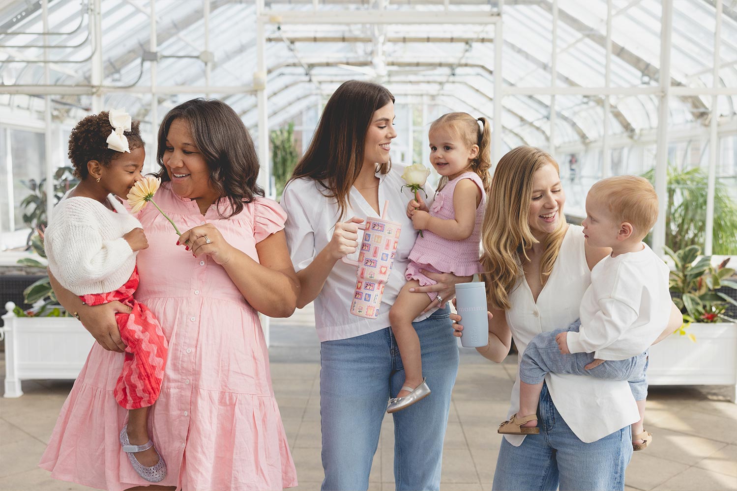 Three women holding children in a bright, glass-roofed space.