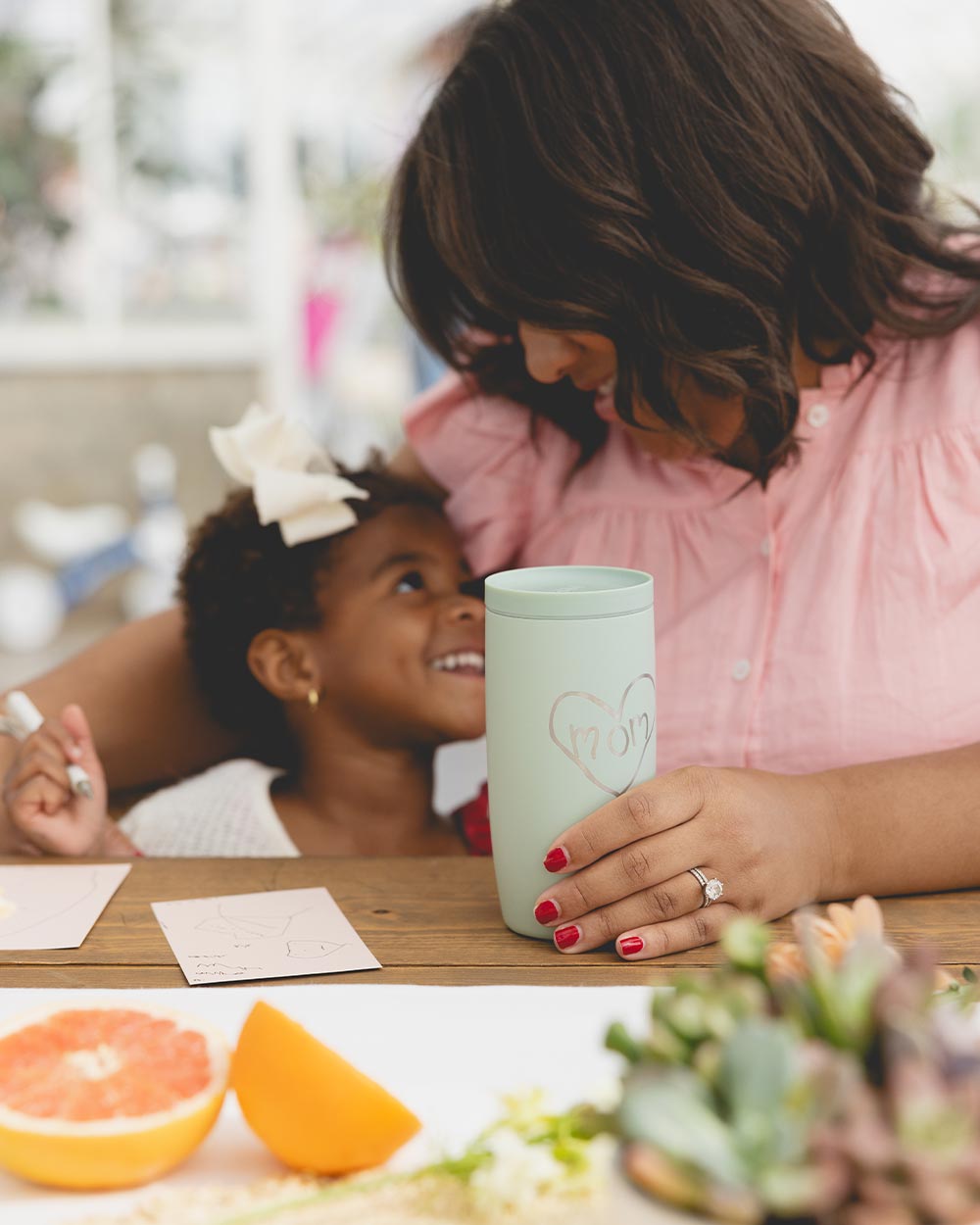 Woman and child smiling at each other, with a mug and fruit on the table.