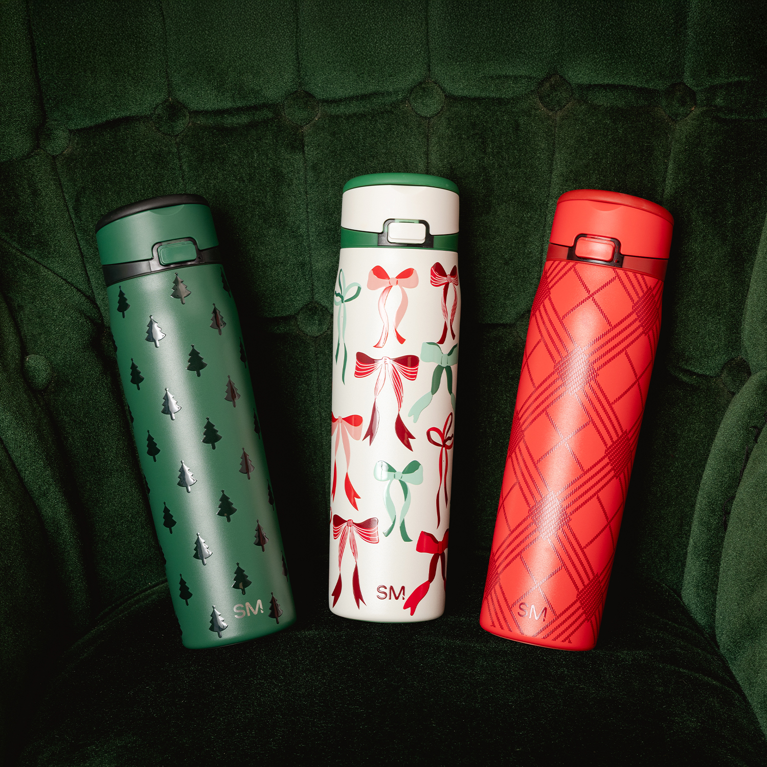 Three festive thermos bottles with holiday designs on a green tufted chair.