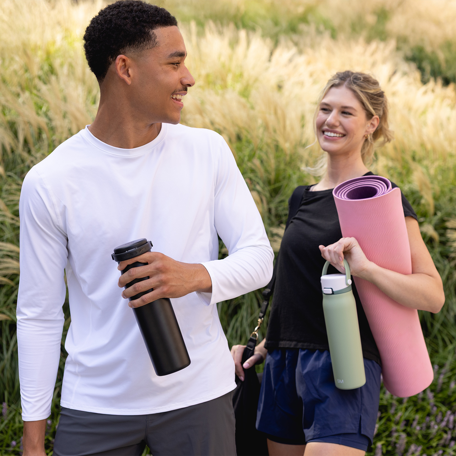 Two people outdoors, holding bottles and a yoga mat, smiling at each other.