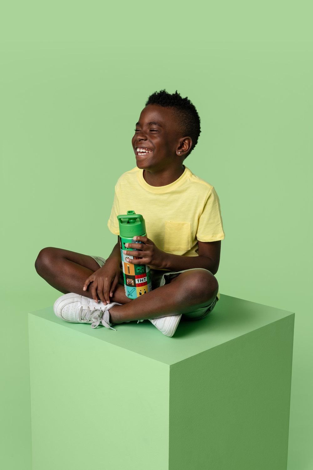 Child in yellow shirt sitting on green cube, holding a colorful bottle, smiling.