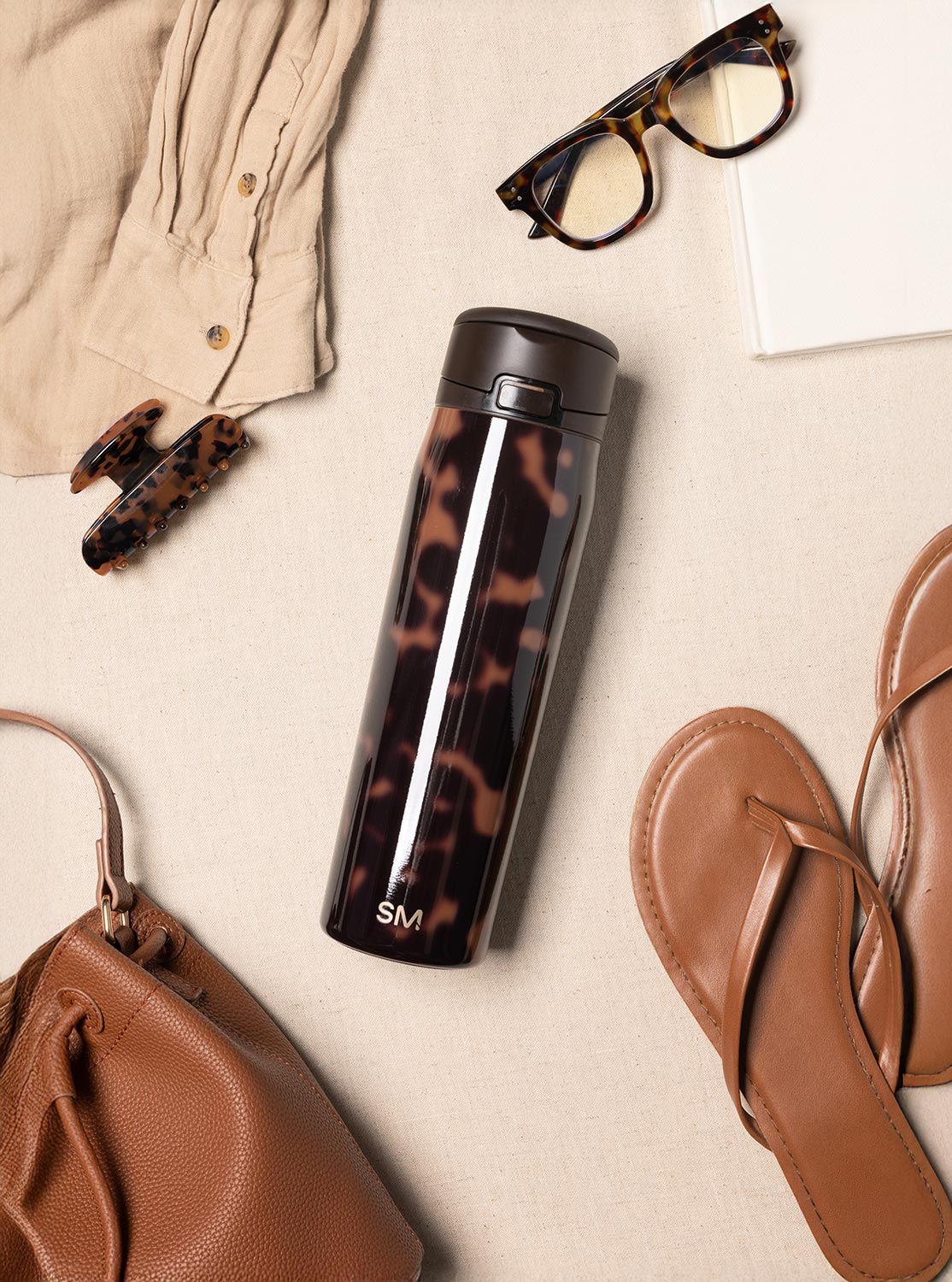 A flat lay of a tortoiseshell travel mug surrounded by matching accessories, a shirt, and sandals.