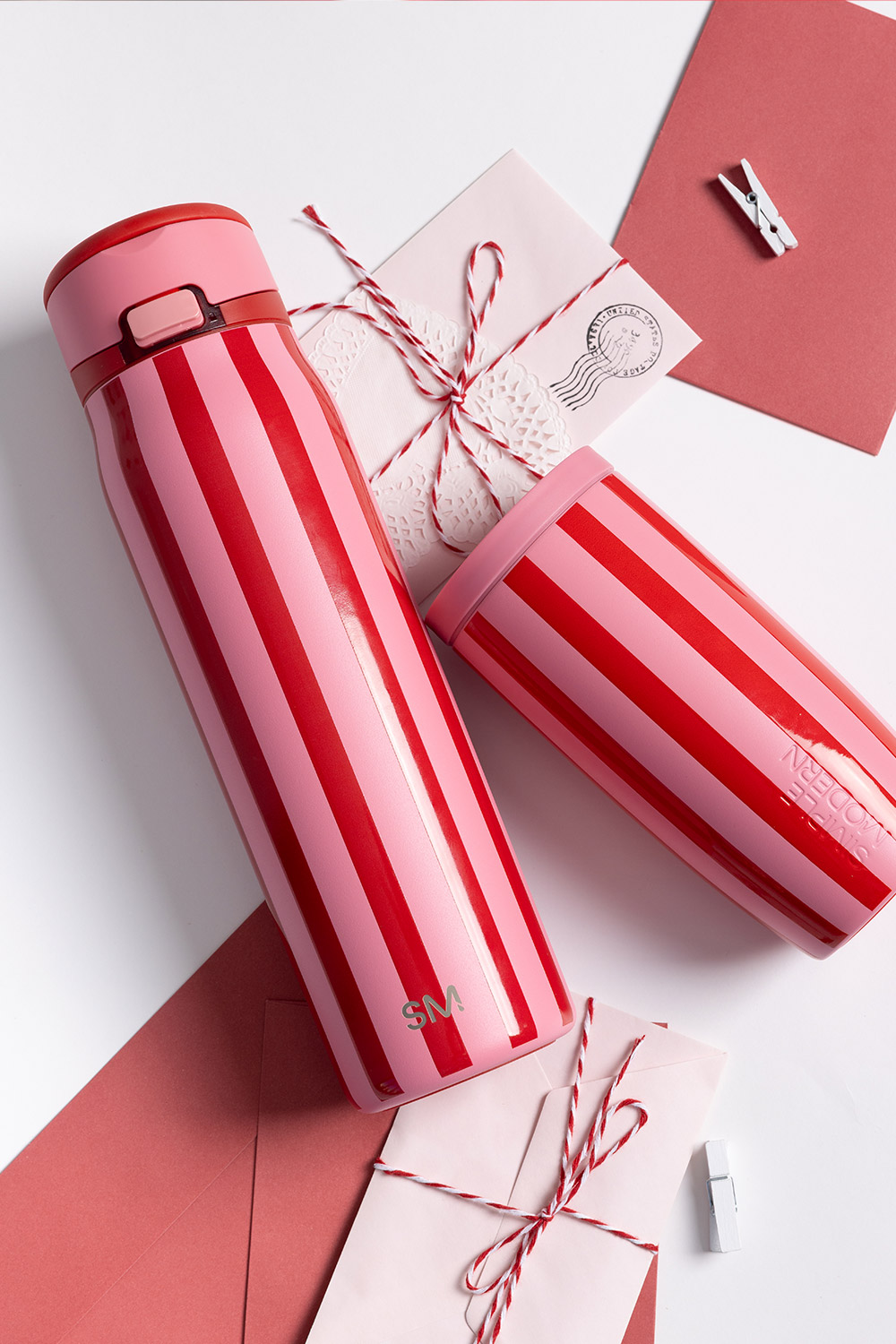 A red and pink striped thermos and tumbler arranged with matching envelopes and string on a white background.