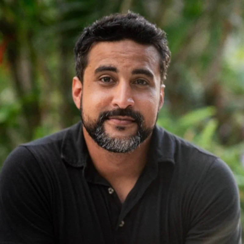 Man with a beard and black shirt, outdoors with greenery background.