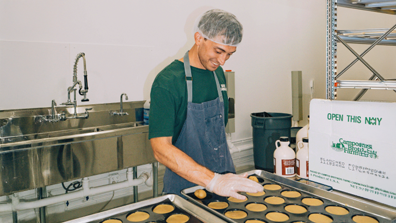Person in apron and hairnet preparing food trays in a kitchen.