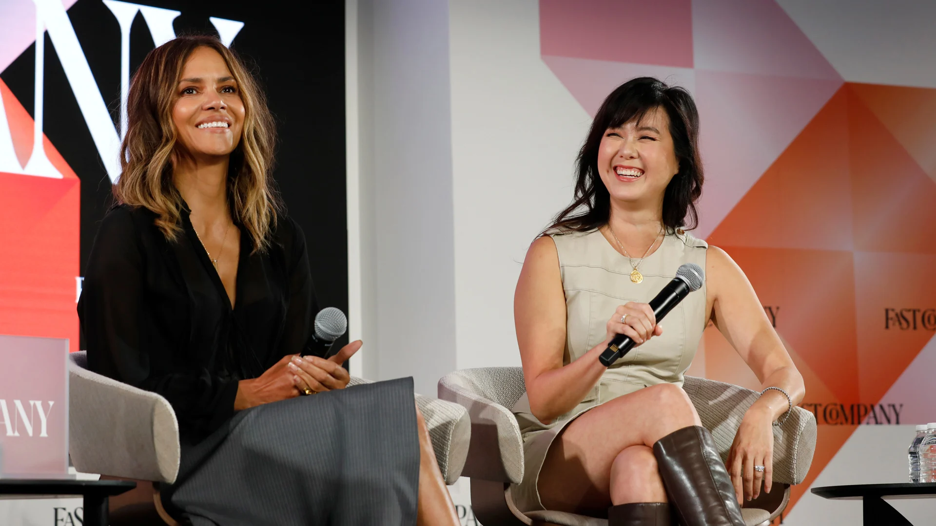 Halle Berry and another woman sit on stage, smiling and holding microphones during a panel discussion.