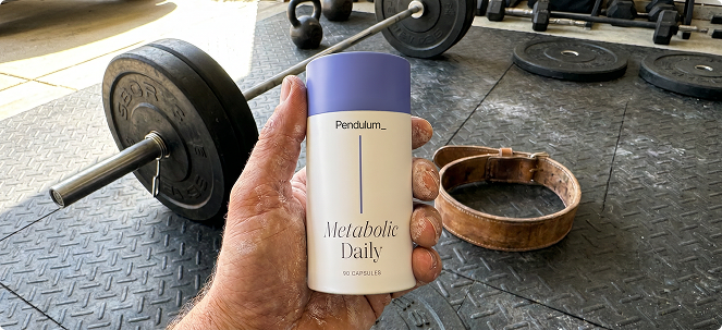 A person's hand holds a bottle of Pendulum Metabolic Daily supplements in a gym setting.