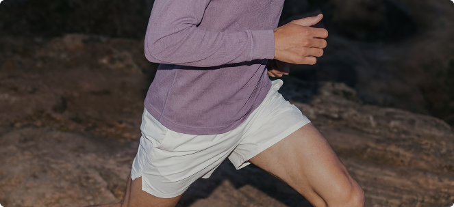 A person in a purple long-sleeved shirt and light-colored shorts running outdoors against a rocky background.