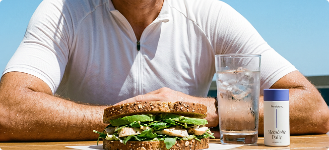 A person in a white shirt sits at a table with a sandwich, a glass of water, and supplements.