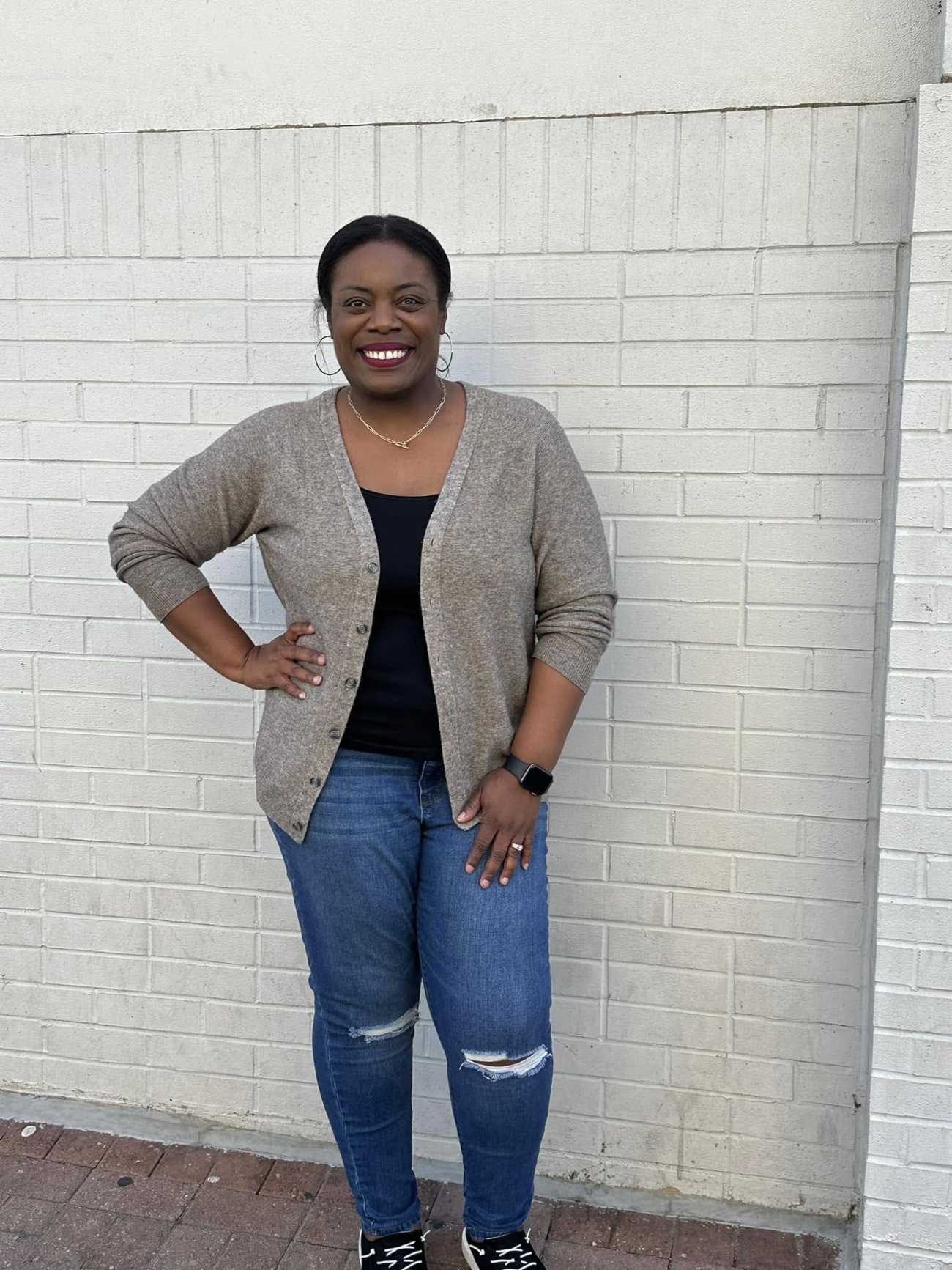 Customer Teri N. standing confidently against a white brick wall while wearing a gray Oliver Charles cardigan over a black top.