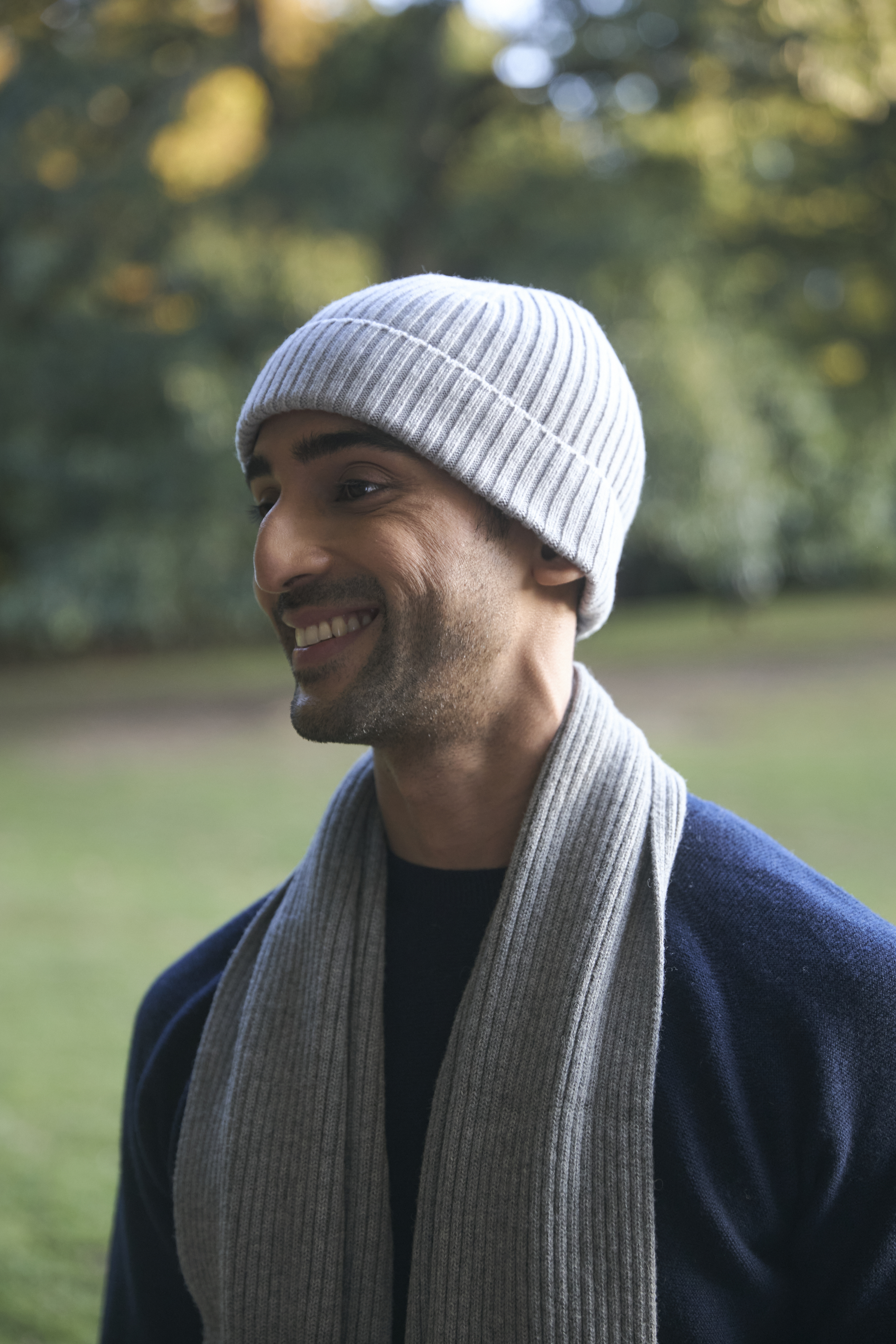 Smiling person wearing a gray beanie and scarf outdoors. 