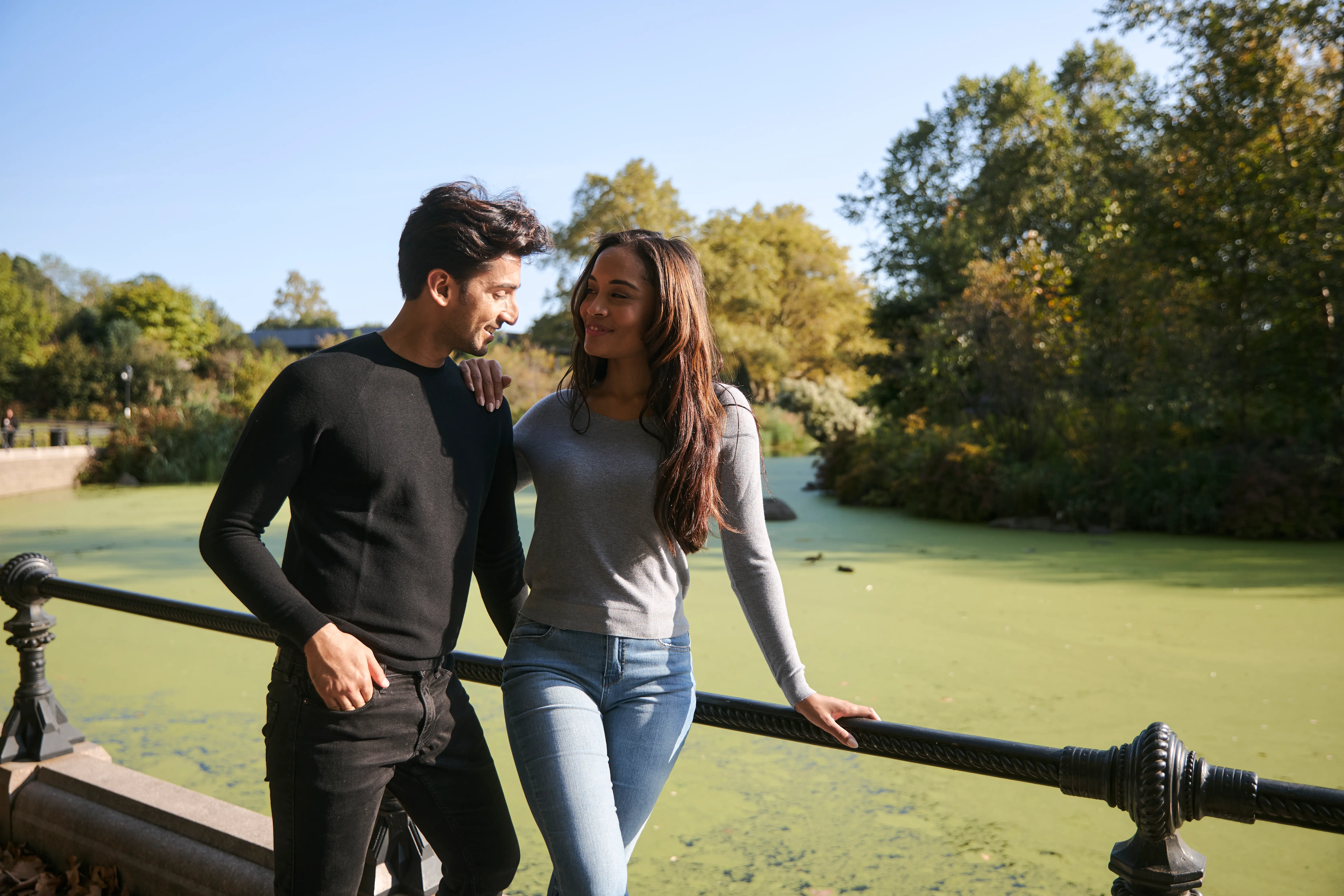 A couple smiling by a pond with green water and trees in the background. 
