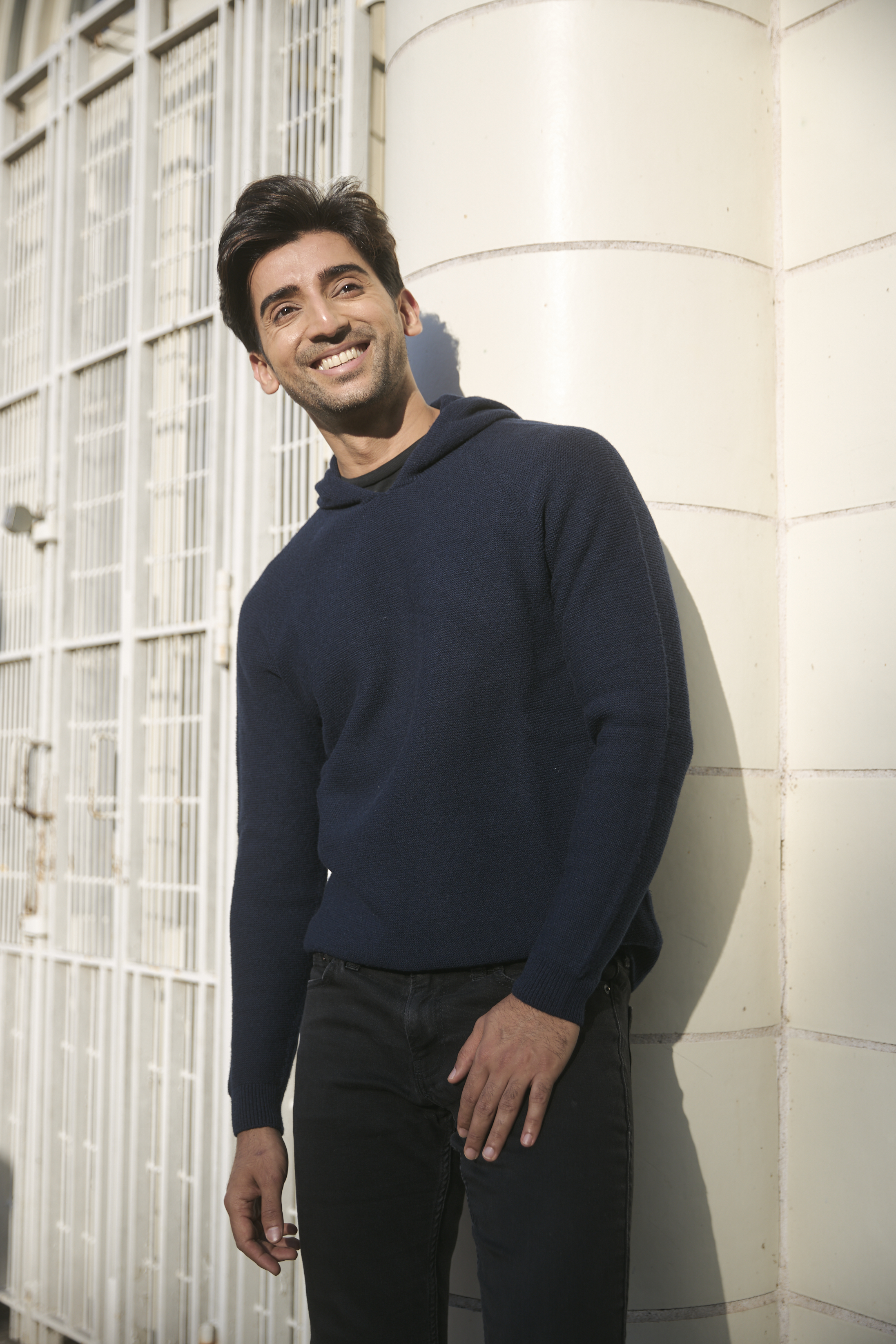 Smiling man in a blue sweater leaning against a light-colored wall. 