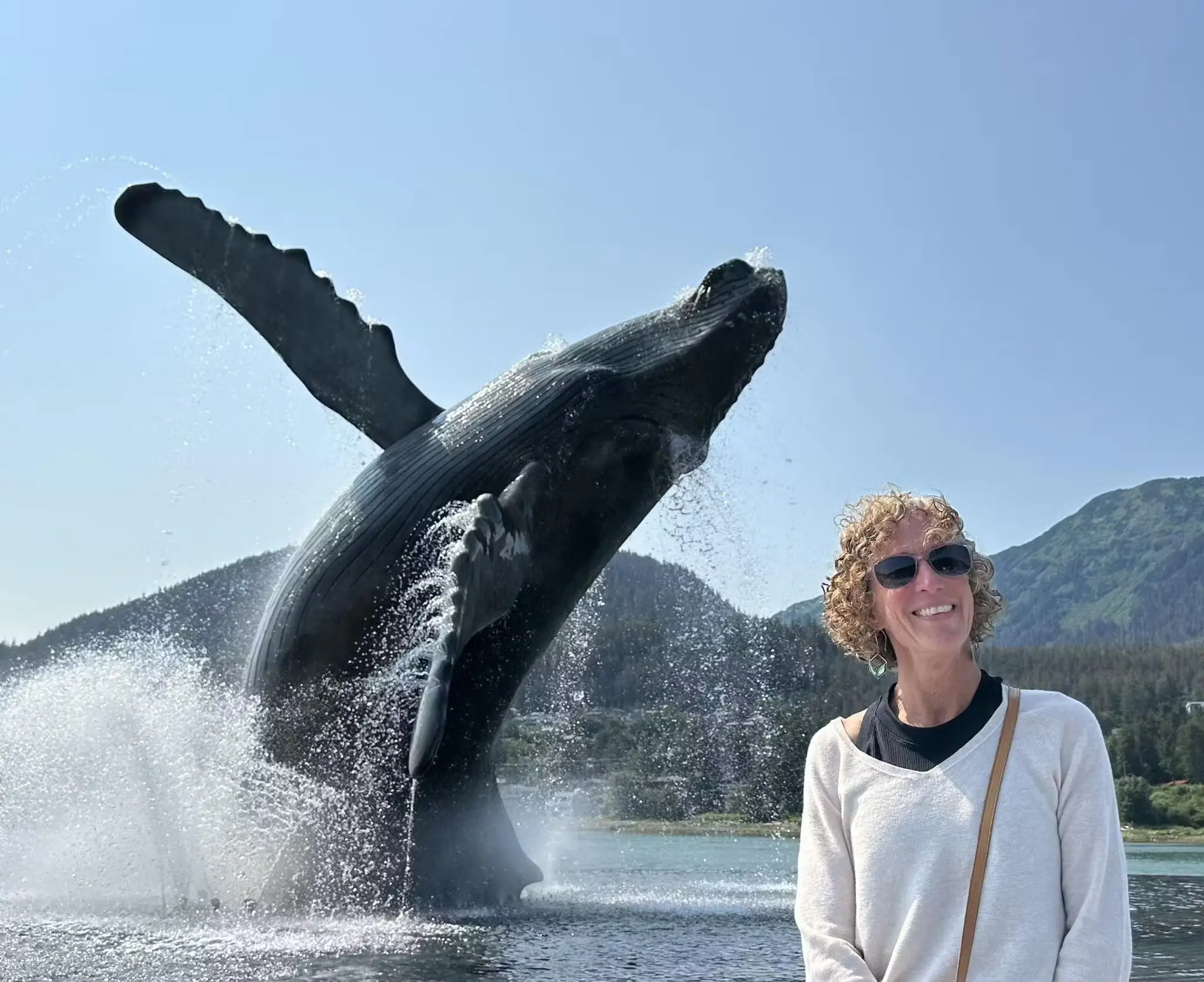 Woman in a beige sweater,  smiling in front of a breaching whale sculpture with mountains in the background.