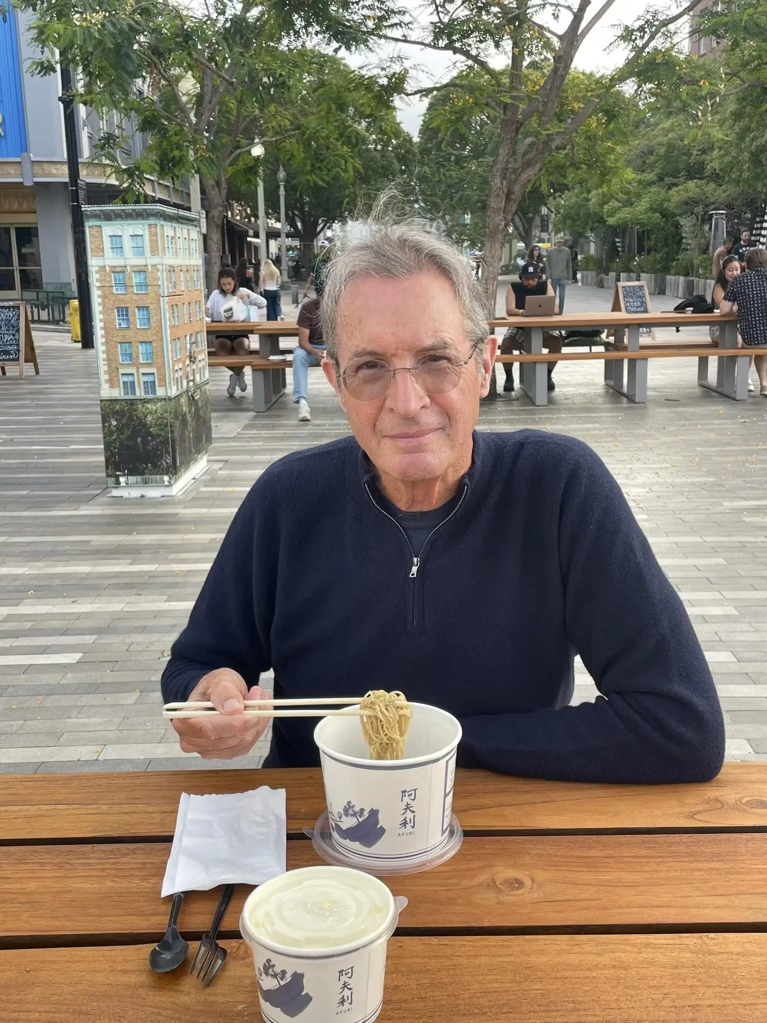 Man in a quarter zip sweater eating noodles with chopsticks at an outdoor table.