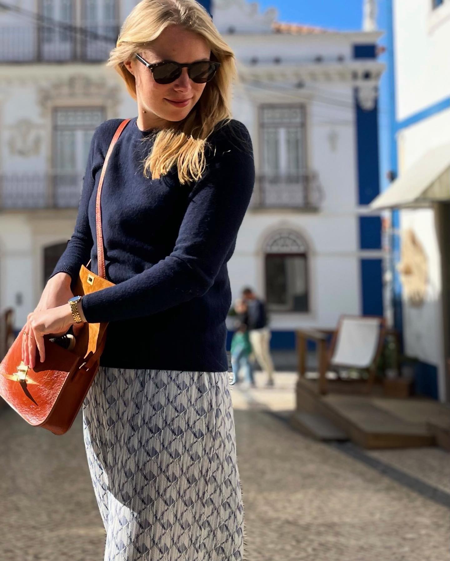 Woman in sunglasses and patterned skirt standing outdoors in a sunny street.
