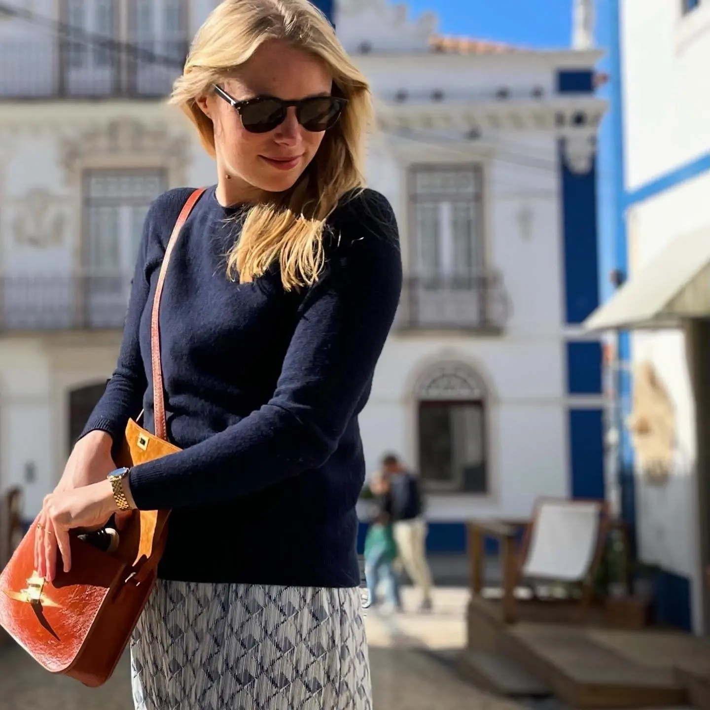 Woman in sunglasses with a brown bag, standing outdoors in a sunny urban setting.