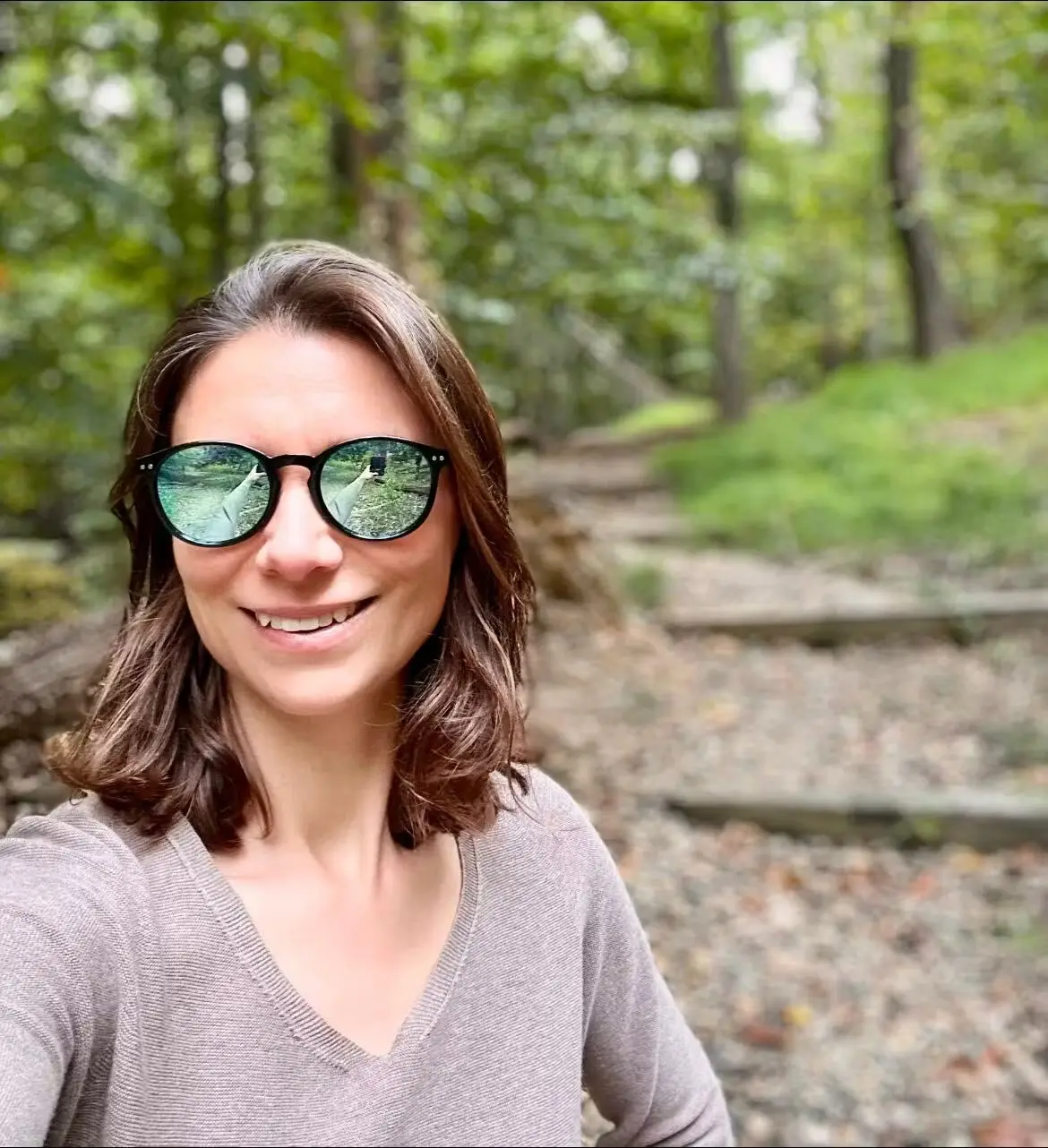 Person wearing sunglasses and a v-neck sweater smiling in a forest setting.