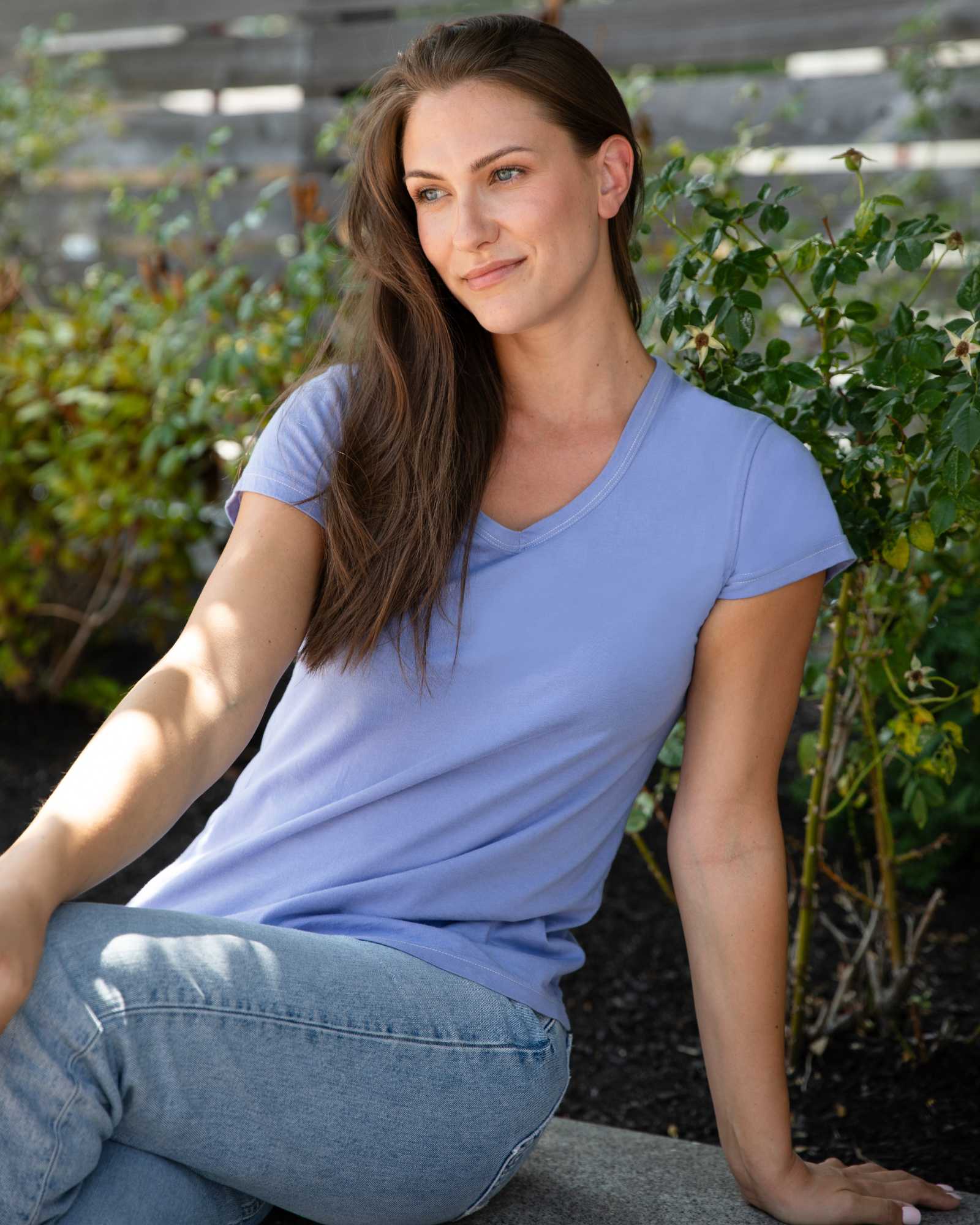 A woman with long brown hair wearing a light blue v-neck t-shirt sits outdoors next to a bush.