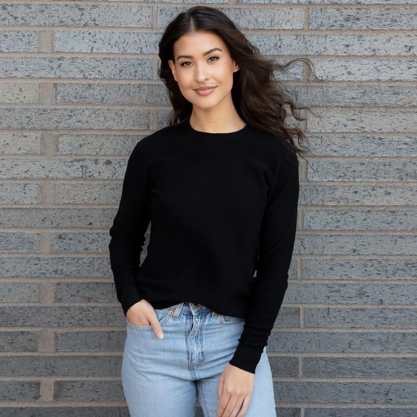 Person in black sweater and jeans standing against a brick wall. 