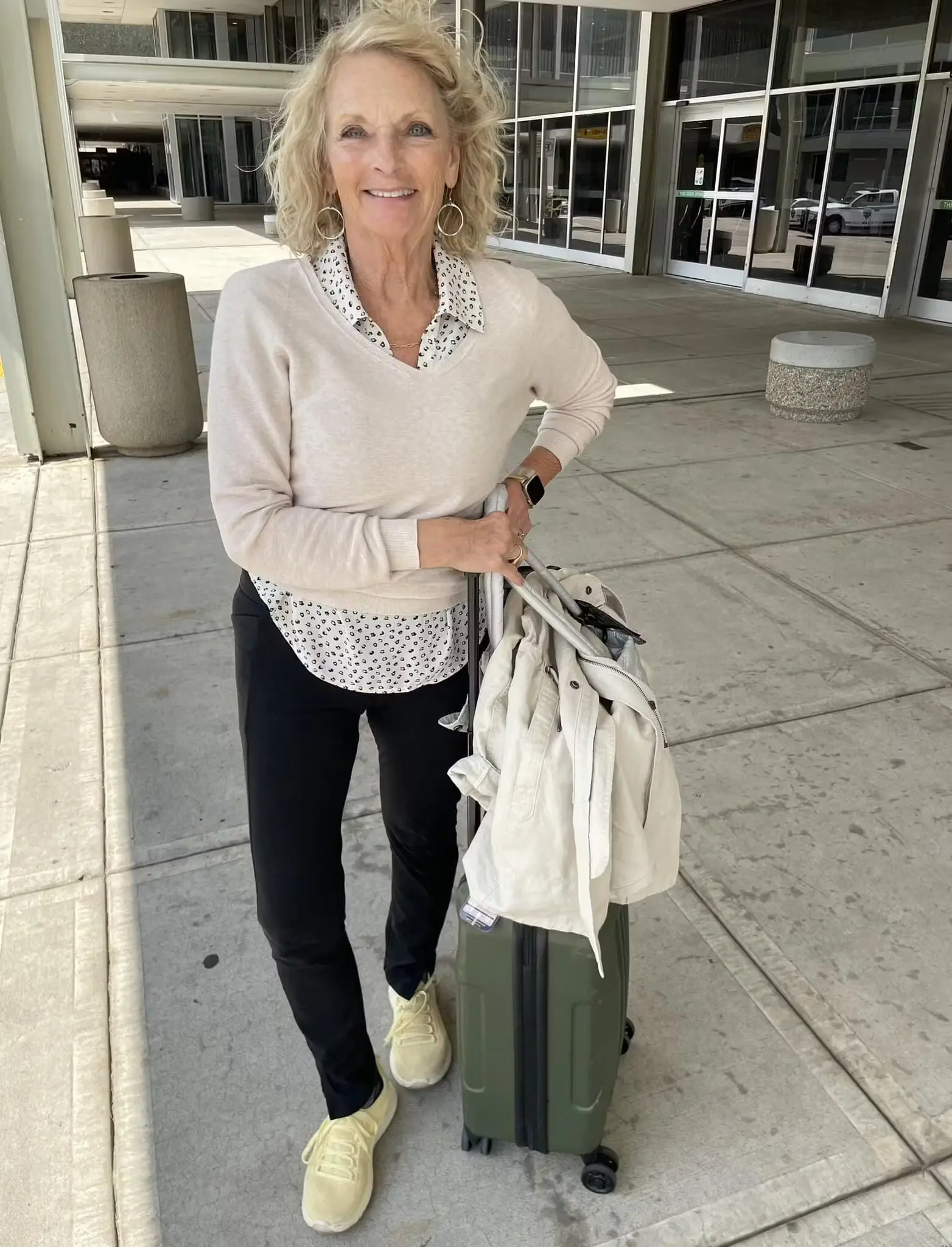 Woman standing outside with a suitcase, wearing a beige sweater and smiling.