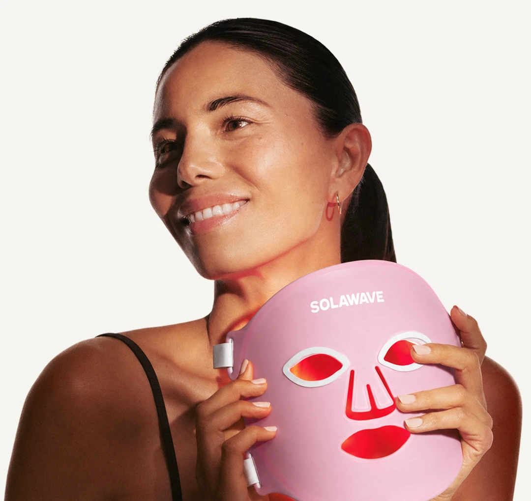 A woman smiles while holding a pink Solawave LED light therapy mask that is glowing red.