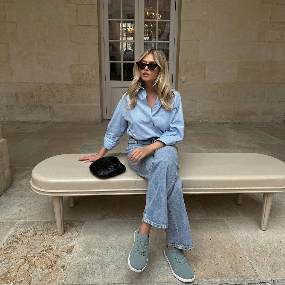 A woman with blonde hair and sunglasses sits on a bench wearing a blue shirt and jeans.