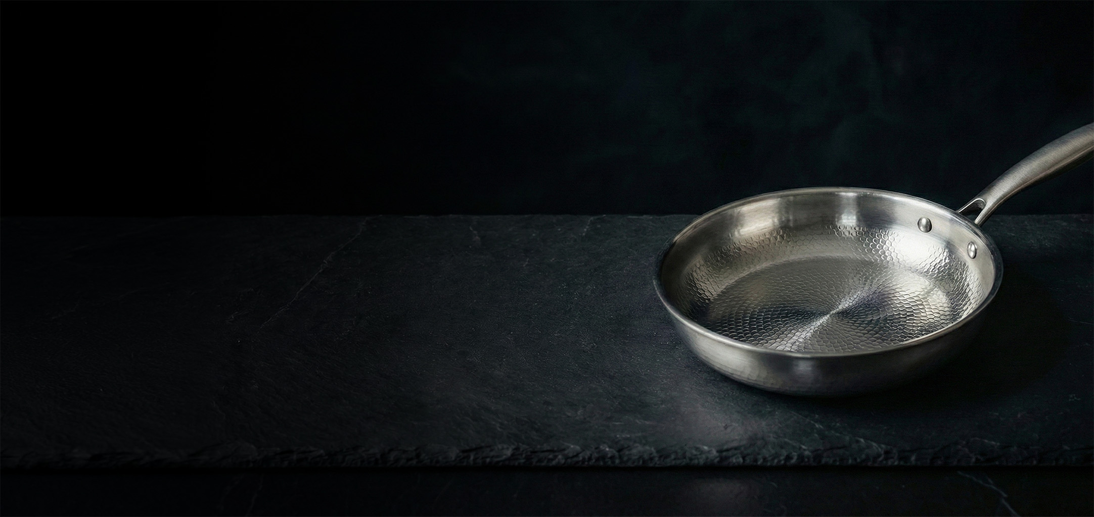 A textured stainless steel frying pan rests on a dark slate surface against a black background.