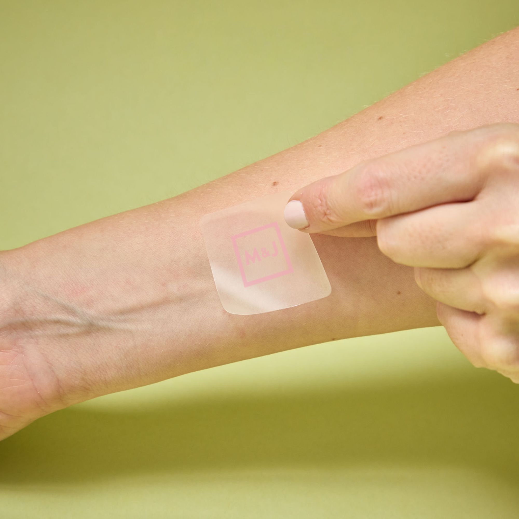 A person applies a clear, temporary tattoo with a pink 'M&J' logo onto their arm.