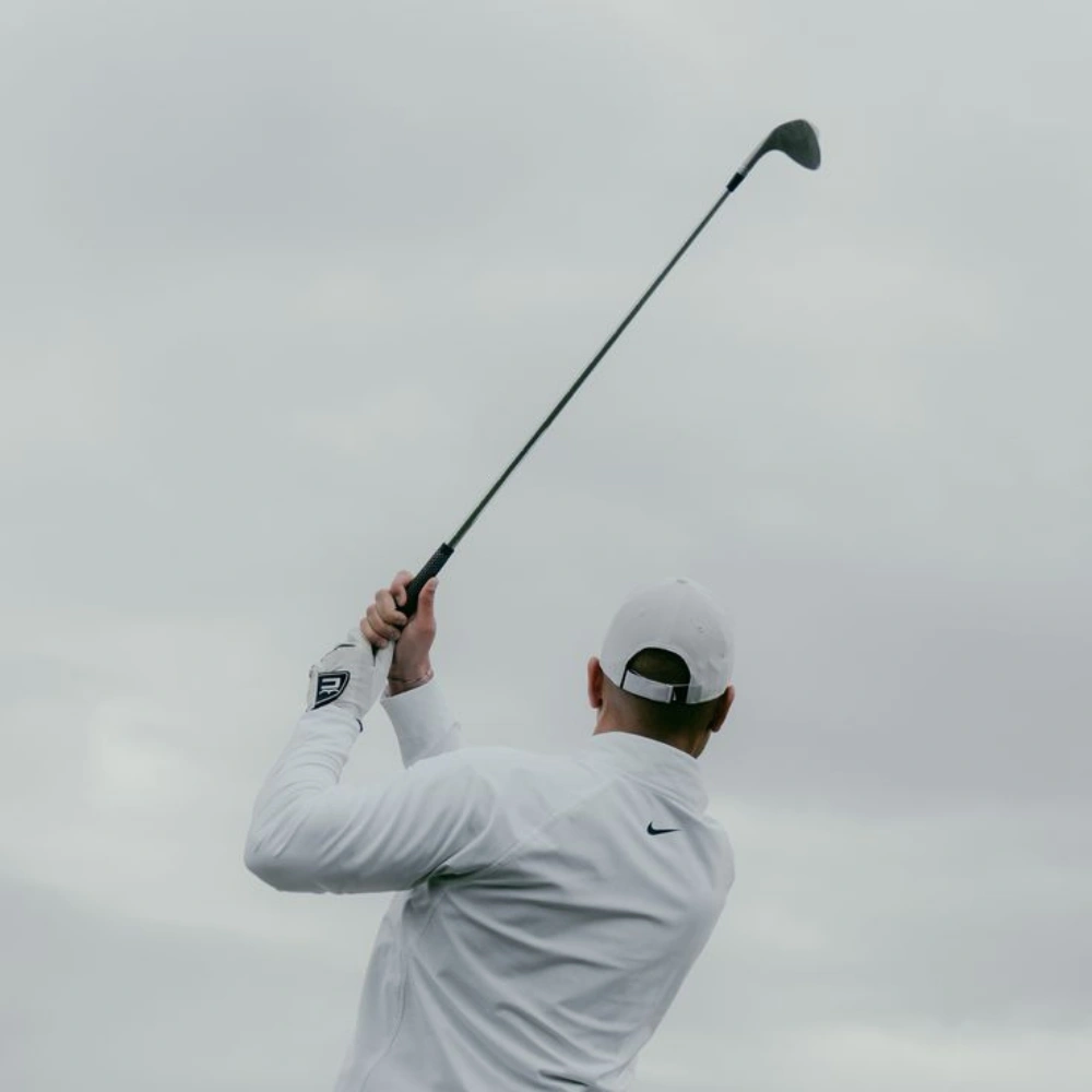 Person swinging a golf club outdoors, wearing white clothing and a cap.