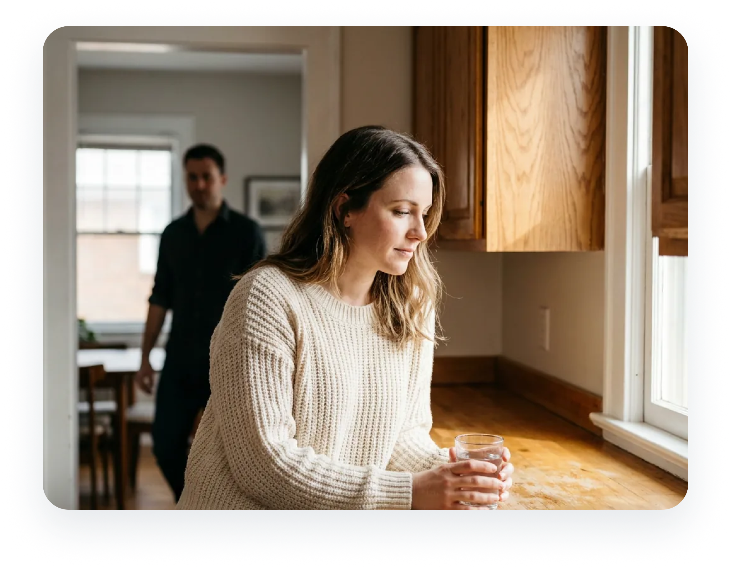 A woman stands in a kitchen holding a glass of water, while a man walks in the background.