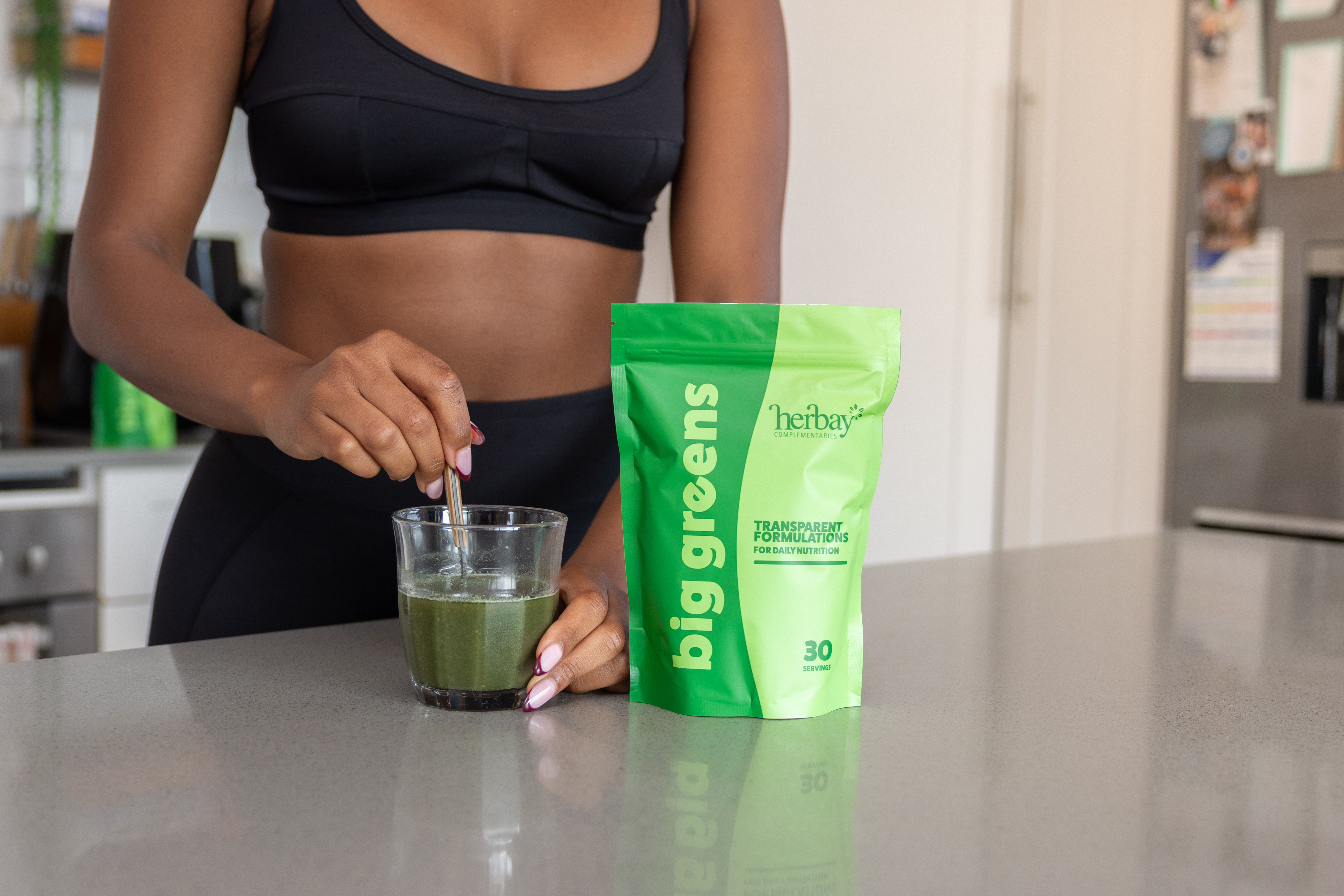 Person stirring green drink next to a green supplement package on a kitchen counter.