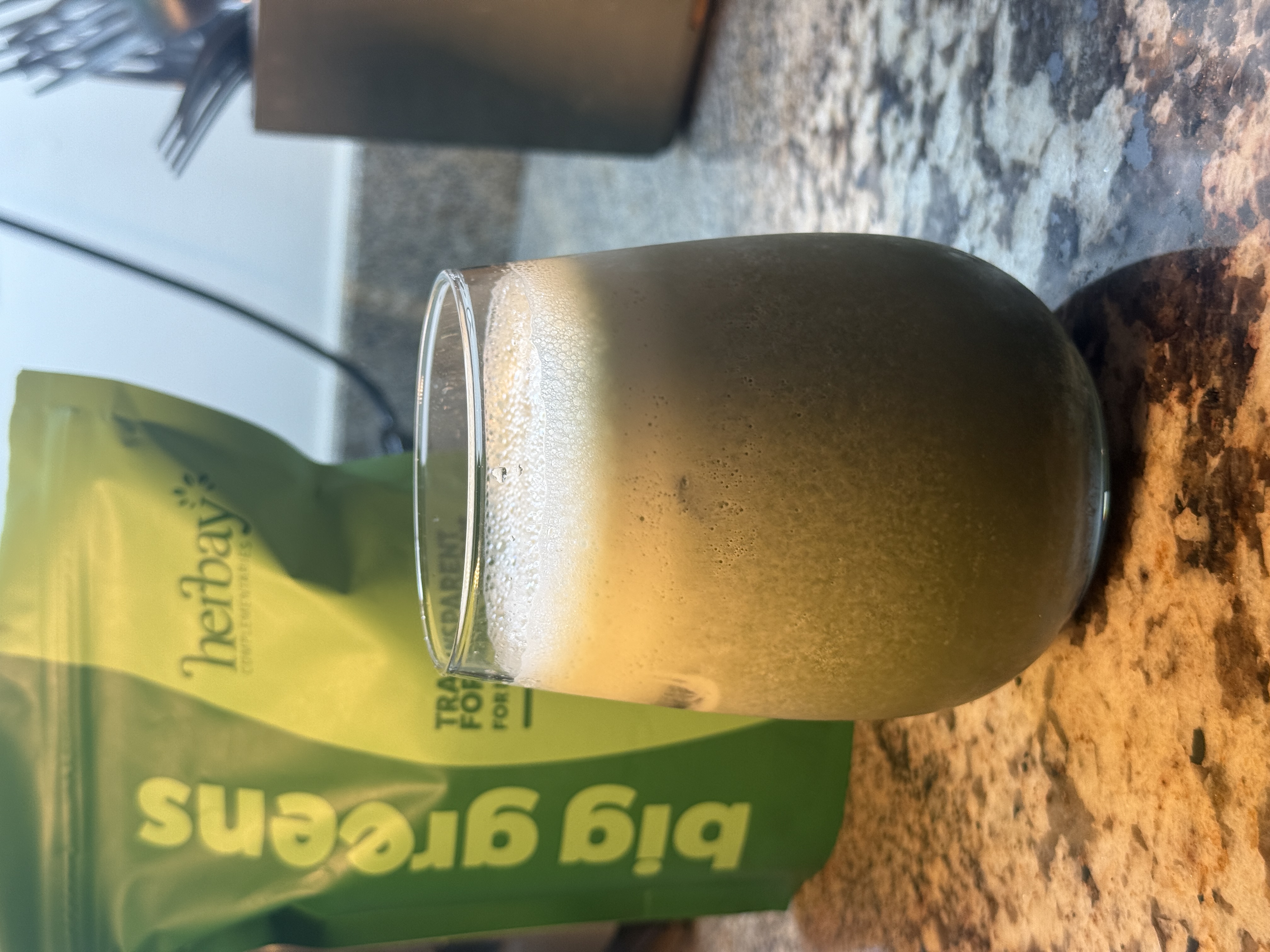 A glass of green smoothie on a kitchen counter with a green powder bag in the background.