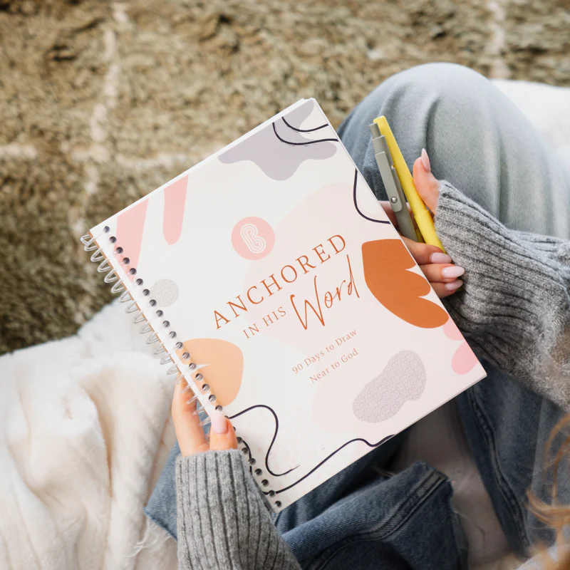 Person holding a journal titled 'Anchored in His Word' on their lap.