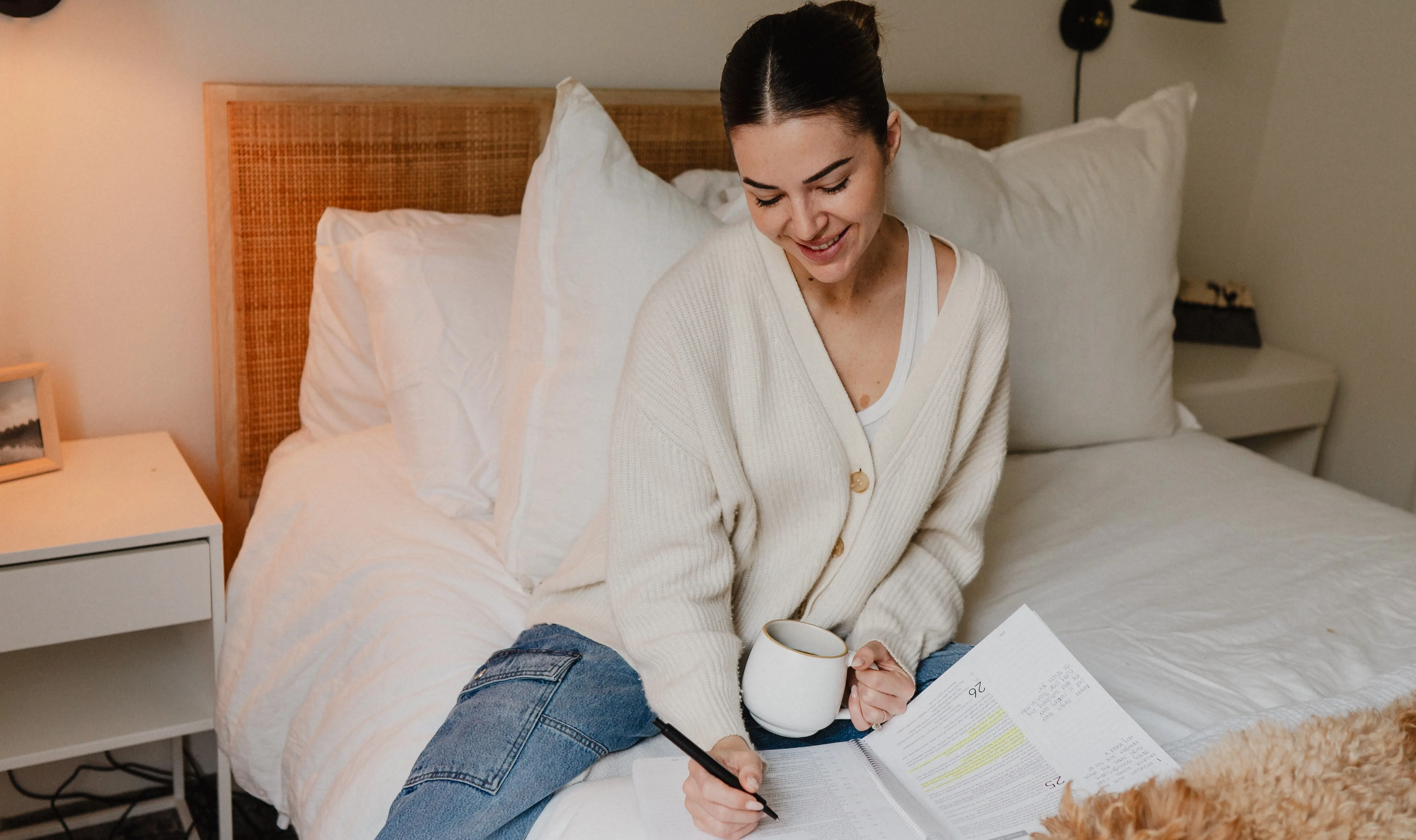 Person sitting on a bed, writing in a notebook, holding a mug.