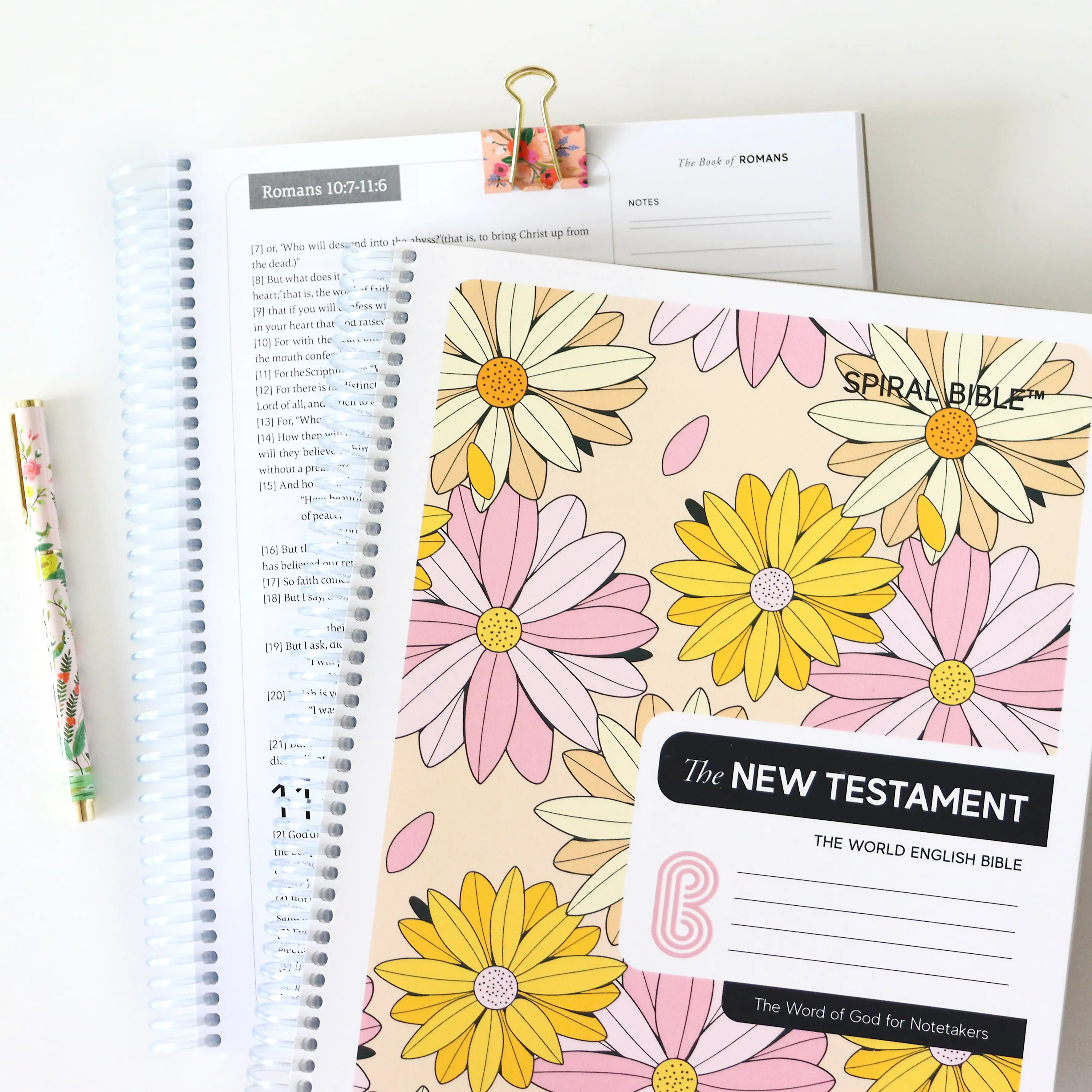 Floral-covered spiral notebook and open book with a floral pen on a white background.