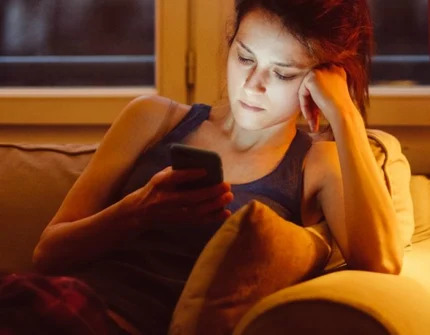 A woman sits on a couch in a dimly lit room, her face illuminated by her phone.