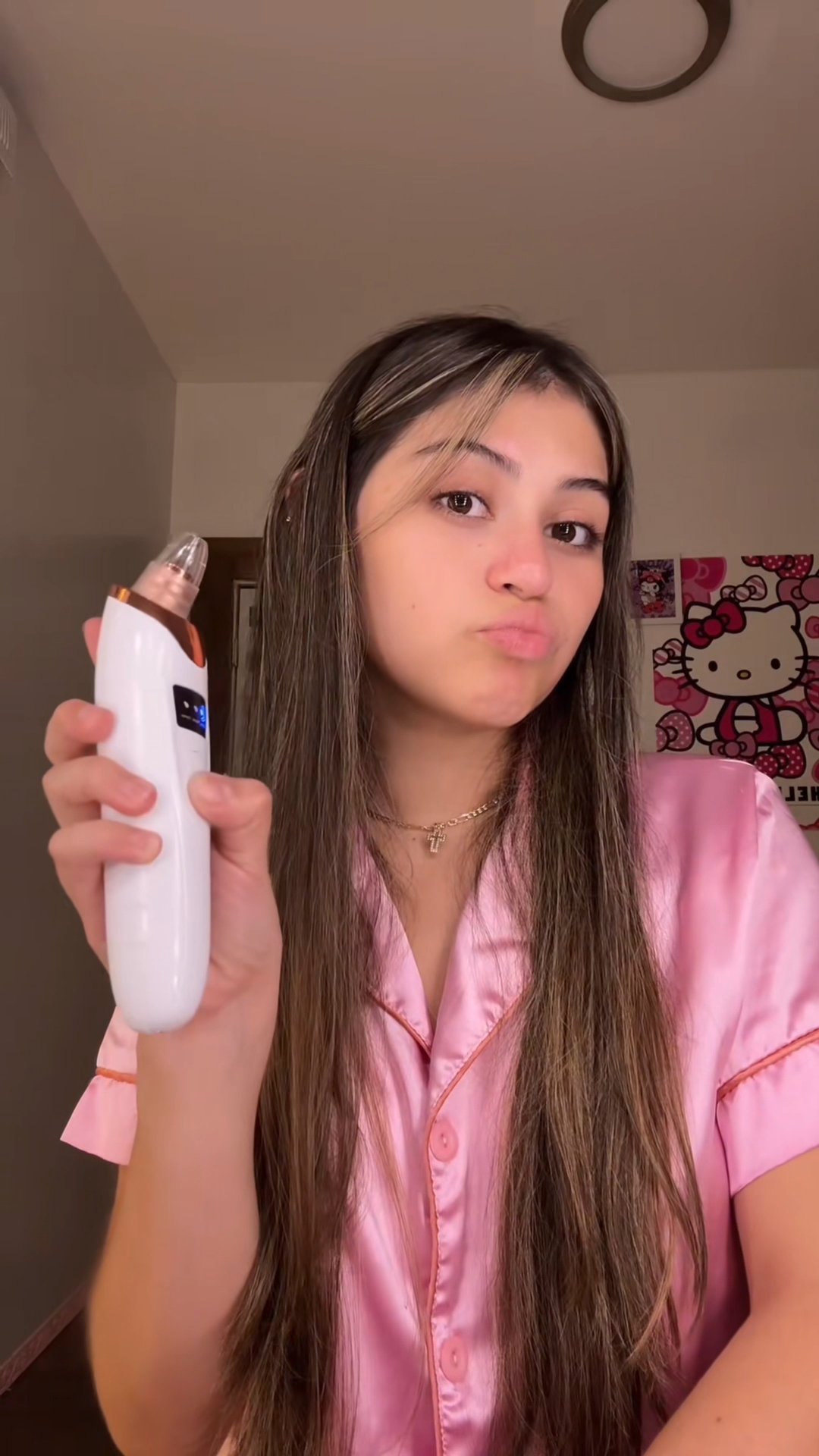 Person in pink pajamas holding a white skincare device, with a cartoon poster in the background.