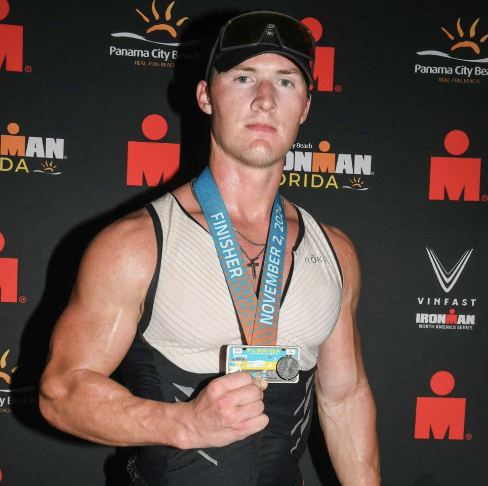 A male athlete poses with his Ironman finisher medal in front of a sponsor backdrop.