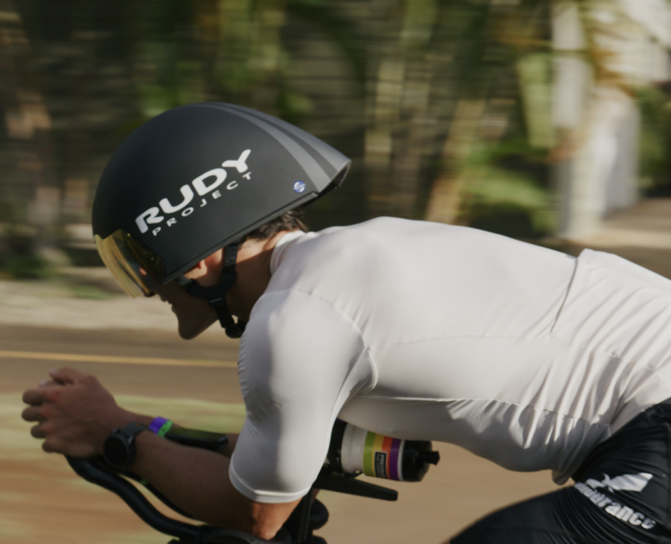 A cyclist in a white jersey and black aero helmet leans forward while riding on a road.