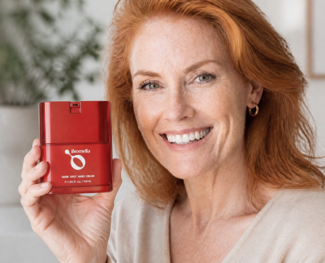 A smiling woman with red hair holds up a red container of Biomella Dark Spot Hand Cream.