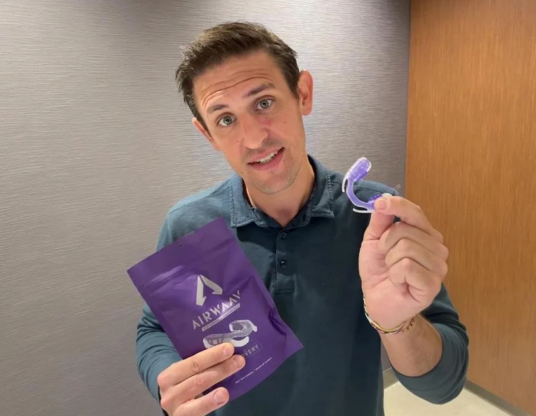 A man in a polo shirt holds up a purple pouch and a purple mouthguard-like device.