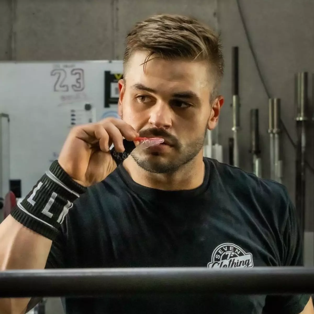 A man in a gym wearing a black t-shirt and wristband holds a mouthguard to his mouth.
