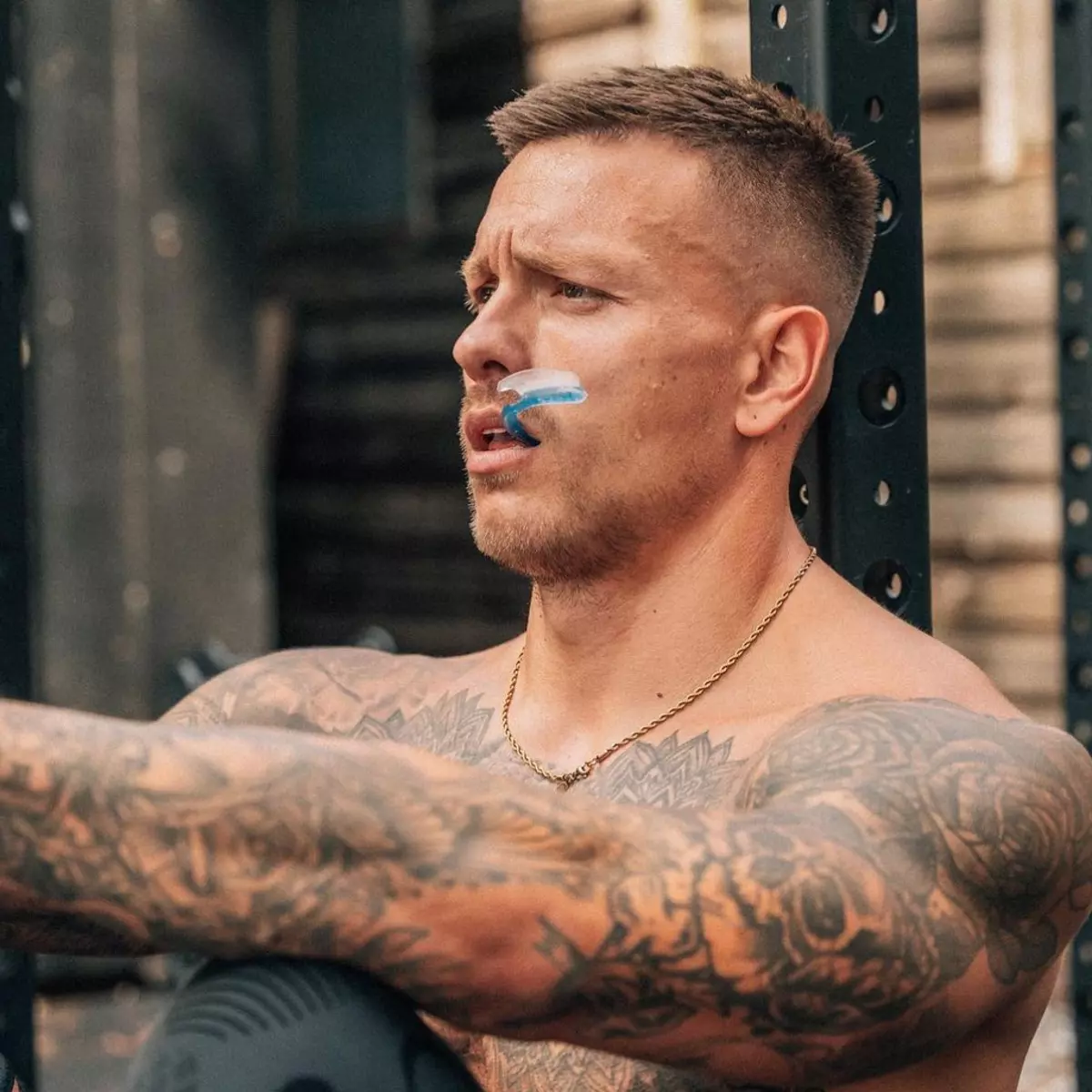 A shirtless, tattooed man wearing a blue and clear mouthguard looks to his right while resting.