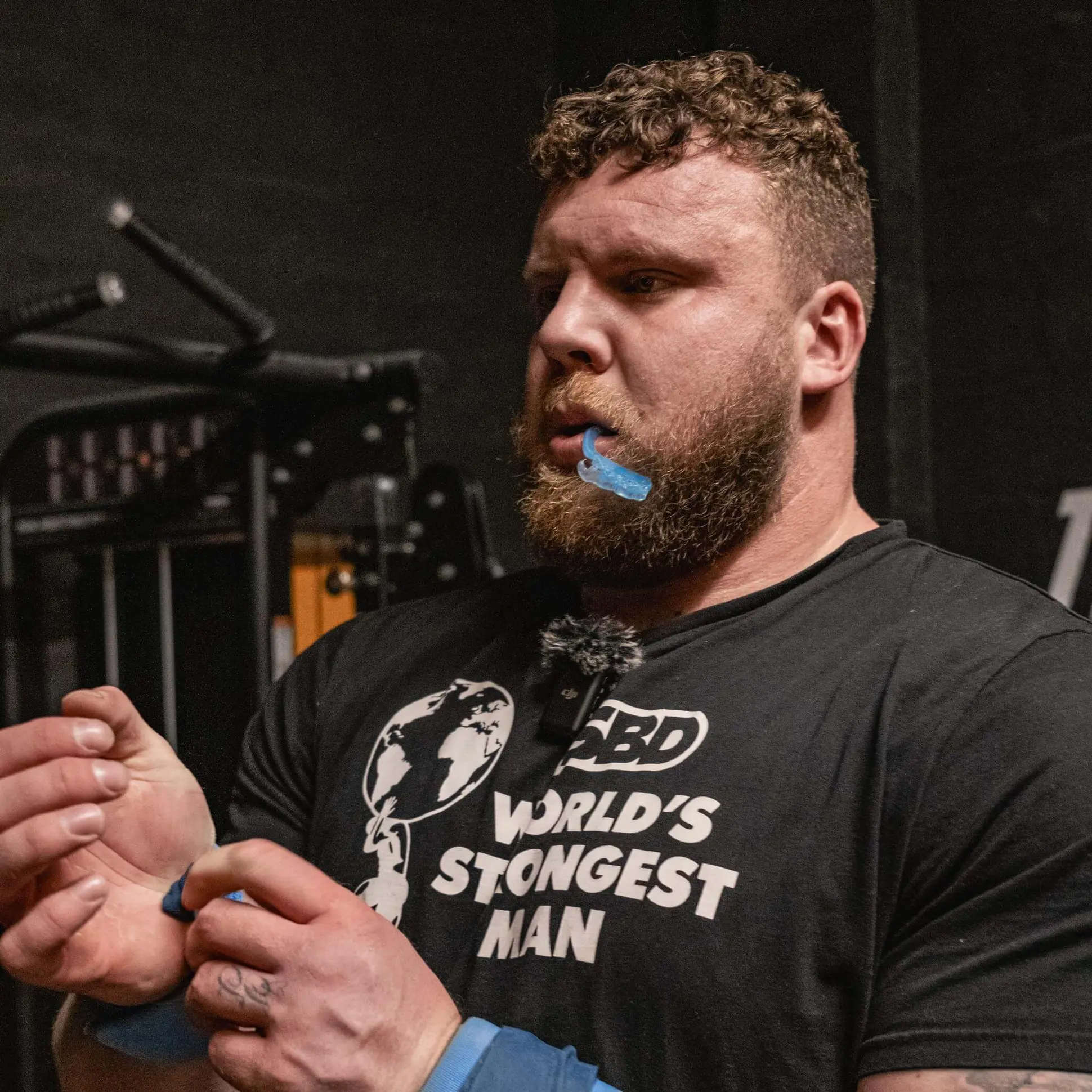 A man in a 'World's Strongest Man' t-shirt with a mouthguard wraps his wrist with blue tape.