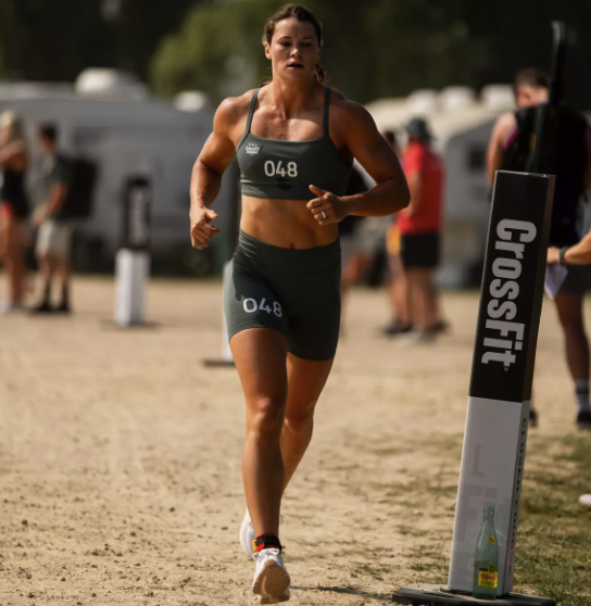 A female athlete wearing bib number 048 runs on a dirt path during a CrossFit event.